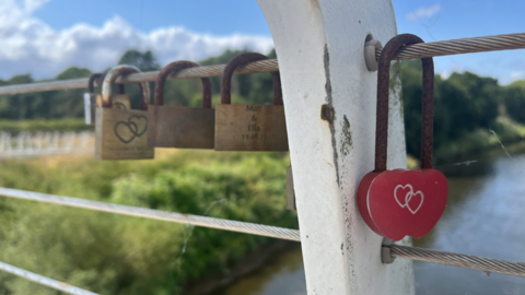 A close-up of padlocks hanging from steel wires on a bridge over a river. The one in clear focus is red and heart shaped, with hearts etched in it. Others are gold with writing or hearts etched on. The padlock clips are rusty. 