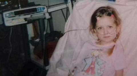 A young girl sits in a hospital bed, wearing a pink nightie with a cartoon fairy design on it. She has tubes attached to her face. She has brown hair and is smiling slightly. Hospital equipment can be seen in the background.