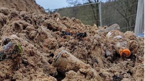 Picture of a mound of sand with lots of rubbish mixed in. In the photo there is broken glass, rusty tines and bottles