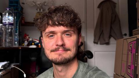 A selfie photograph of Ben Cole Edwards, who is 30 years old, has brown curly hair, a brown beard, and blue eyes. Behind him, a coat can be seen on the hook on his back door, and a plastic water bottle can be seen on a desk behind him. 