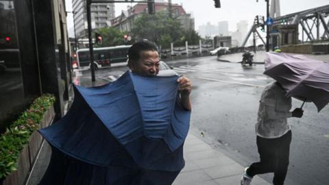 A man, looking uncomfortable, struggles with his umbrella on a Shanghai street. Another person to his side is having the same issue.