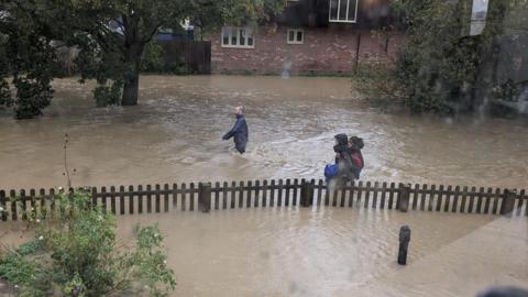 Three people wade through high flood waters in Debenham following Storm Babet in 2023. The water reaches their waists and they wear coats. A fence can be seen protruding slightly above the water and homes can be seen in the background. 