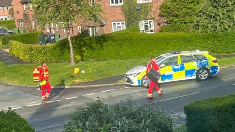 Two paramedics wearing red uniforms with yellow stripes are seen walking along a road next to a West Mercia Police branded vehicle.