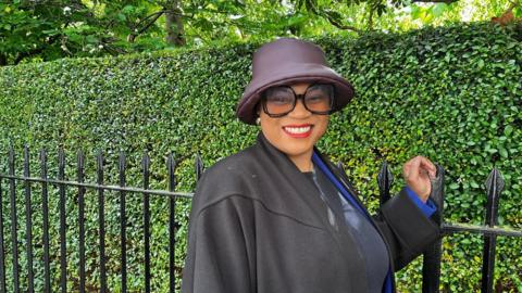 Woman wearing a sunglasses and hat, smiling against a backdrop of green bushes and trees.