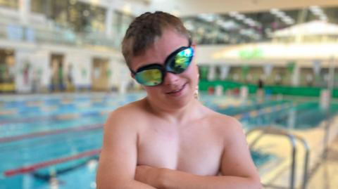 Sam Craddock wearing reflective swim goggles stands beside an indoor swimming pool with arms crossed. He is positioned in the foreground near the pool edge. The background shows swimming lanes, poolside railings, and a spacious aquatic center interior with bright lighting and blurred architectural details.