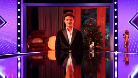 Lando Norris addresses the Sports Personality of the Year Awards via video link. He is on a screen wearing a suit jacket and in front of a Christmas Tree. The shot is of the screen on a purple lit stage with a shiny floor and the presenter looking at him to the right of the shot.