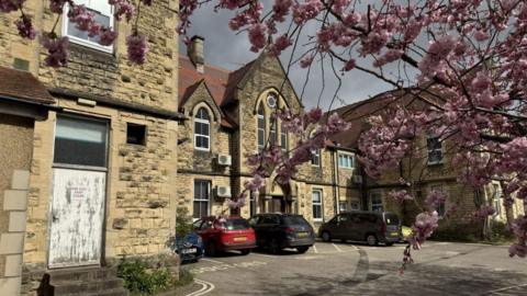 The exterior of the hospital. It is a yellow brick building with Victorian features. Cars are parked in bays outside the front of the hospital, with a large blossom tree to the side of it.