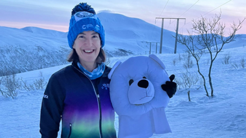 A lady standing in a snowy landscape, holding a fancy dress polar bear head and wearing a blue bobble hat. She is smiling towards the camera and wearing a purple and dark blue jacket. The sky is a pale pink in the background. 