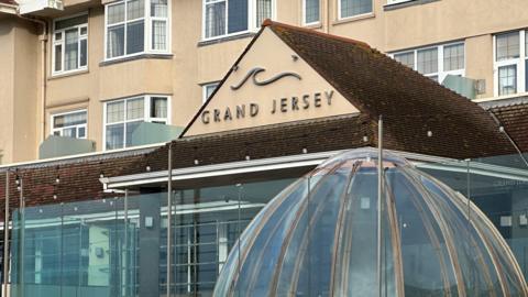 The front of the Grand Hotel in Jersey. The building has a beige facade with multiple windows, and the sign is mounted on a triangular section of the roof. Below the sign, there is a glass structure with a dome-like shape.