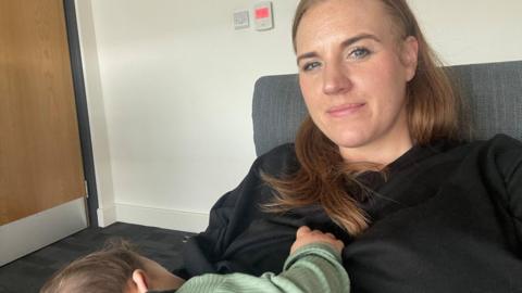 Lauren Somerville. She has long brown hair and is wearing a black top. She is sitting on a chair and her young son is on her knee.