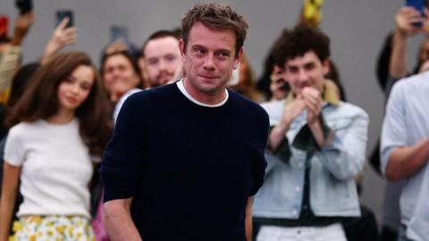 A man wearing a navy jumper and white top, he has dark hair