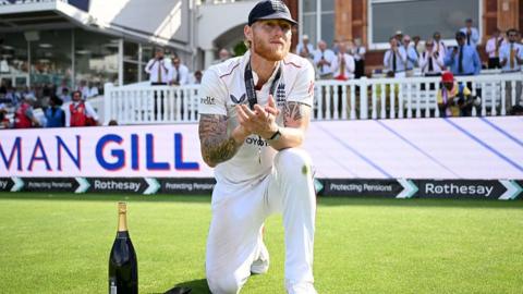 Ben Stokes stands on one knee, applauding, with champagne beside him in a dark bottle