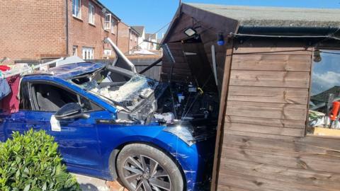 A wide shot of the Blue Skoda crashed into the garden shed.