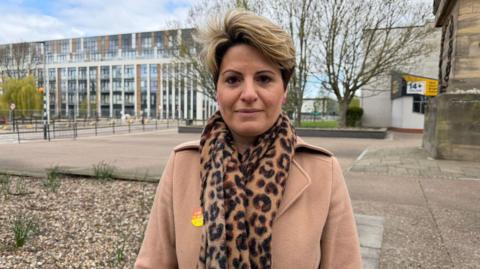 Emma has short hair and wears a beige wool coat and animal print scarf with a labour sticker on her coat.
