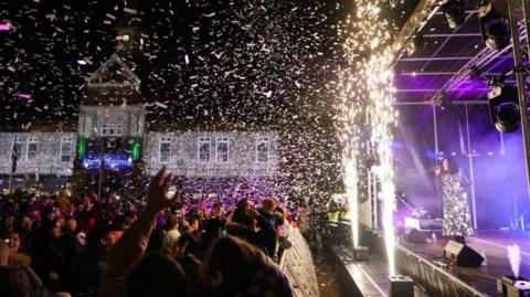 A large crowd of people is standing behind a barrier watching a woman sing into a microphone on a stage. Sparks shoot upwards out of machines at the front of the stage. Confetti is falling onto the crowd from above. Security guards watch the crowd from the front corner of the stage. 