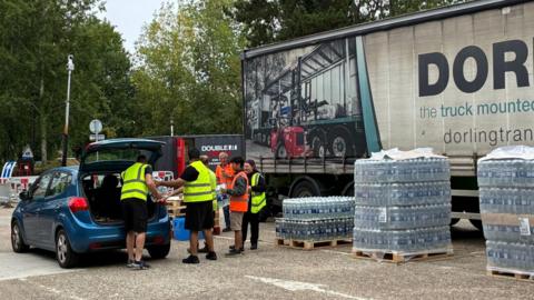 People from South East Water putting bottles of water from the water station into a blue car. There are five people in the image, and they are all wearing high-vis vests.
