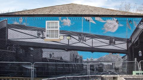 A side of a building with a mural painted on it. The mural shows blue sky and a bridge over a railway line with trains on the tracks. Buildings and walls have been painted alongside the tracks.