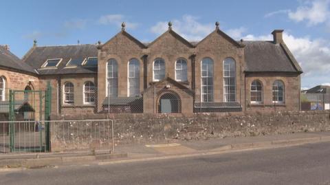 The school is a Victoria era building. There are three gable ends along one side of the school with long windows that are arch shaped at the top. There is a long brick wall separating the school grounds from a pavement and a road.