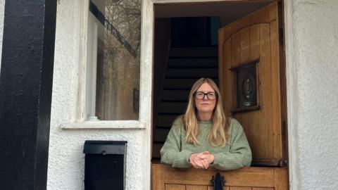 A blonde woman with glasses stands at a stable-style half door at a white and black painted house.