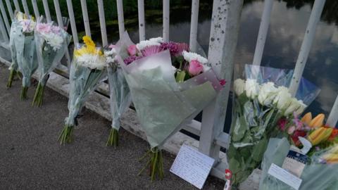 Bouquets of flower tributes are left on the floor leaning up against bridge railings. A note can be seen next to one of them.