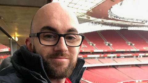 Gregory Dervin smiling while on a tour of Arsenal's Emirates Stadium. He has a beard and shaved head and is wearing glasses and a black coat. Behind him are row upon row of empty red seats.