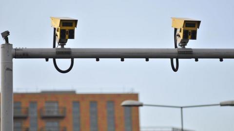 Two average speed cameras installed on a post over a road, with apartment blocks in the background 