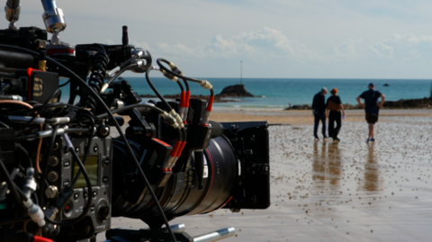 Close-up of a professional film camera positioned on a beach, sharply focused against a blurred coastal background. Three people walk near the water's edge across wet sand reflecting the sky, while calm blue water and a small rocky structure sit along the horizon. 