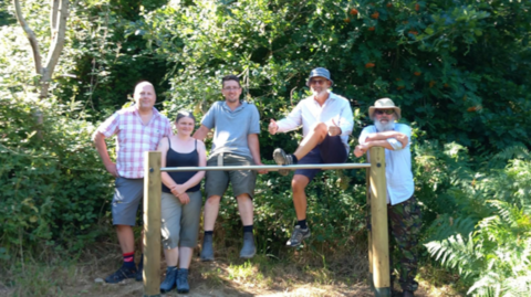 Five people standing behind a new fitness bar in a woodland. The bar is supported by two wooden posts and there is shrubbery around it. There are also a number of trees behind the people.