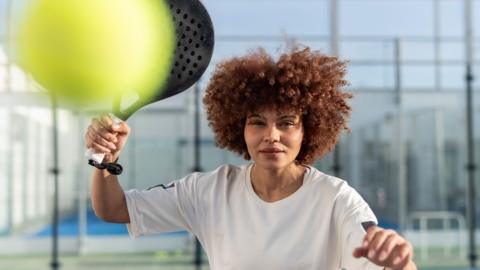 A woman holding a padel racquet about to hit a ball.