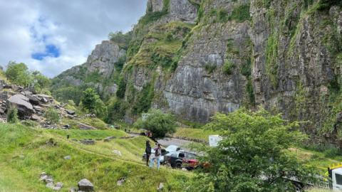 Cheddar Gorge in Somerset with cars parked and the impressive cliffs visible above a bank of trees, grass and rocks