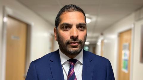 Mr Russell is wearing a dark blue suit, white shirt and purple, yellow and blue tie. He has a closely shaven beard, dark hair and is standing in the hospital corridor.