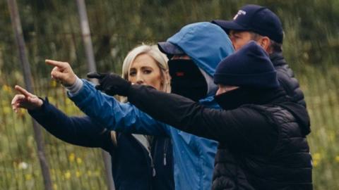 Carla Lockhart standing beside four men. They are all pointing their fingers with their arms in the air.