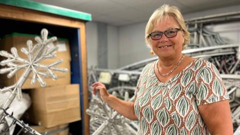 A lady with short blonde hair and glasses wearing an orange and green patterned t-shirt in front of a background of snowflake Christmas lights in a storage unit