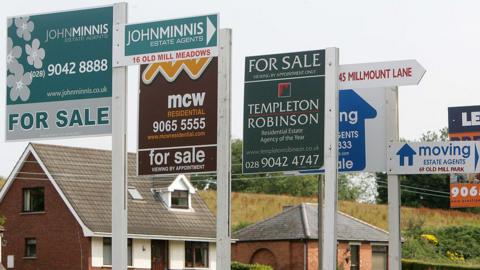 A row of 'For Sale' signs in front of two houses. 