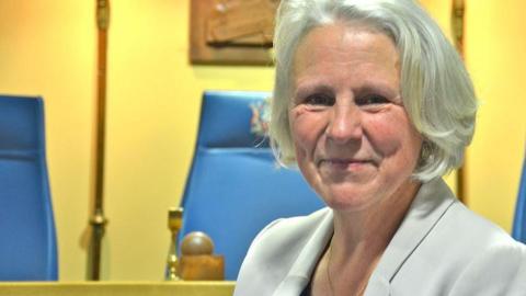 Rossendale Council leader Alyson Barnes sitting in council chambers with grey bobbed hair wearing a light grey suit jacket. She is smiling.  