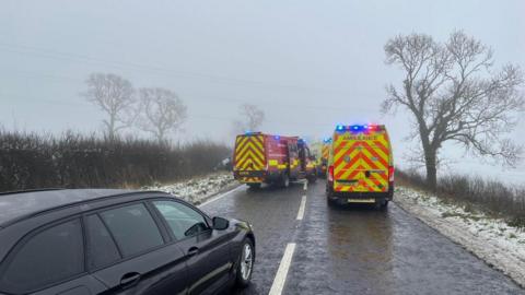 Two ambulances and a fire van are blocking the road. A car can be seen in the ditch to the left of the road. 