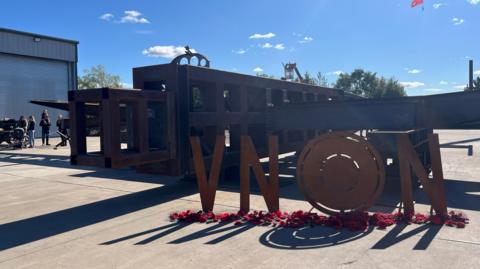 Part of the large metal structure lies on the floor with metal letters V N N and a row of poppies underneath.