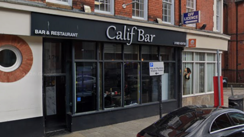 General view of a black shop front with the words Calif Bar in white. A car is parked outside, next to a sign that gives parking restrictions on the street.