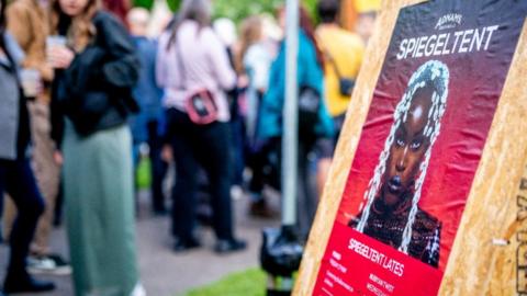 A group of people have gathered at the Norfolk and Norwich Festival in Chapelfield Gardens. A red poster says SPIEGELTENT on a board. 