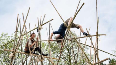 Two performers are balanced on a series of interlinked bamboo poles. One of the performers is upside down with his back arched, the other is sat watching, perched on poles nearby.