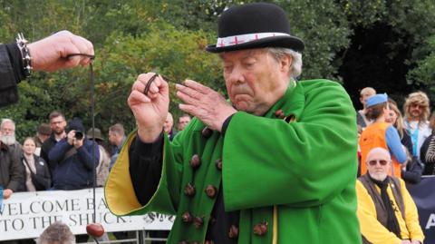 David Jakins is dressed in green and adorned with conkers. He is focusing on a game of conkers he is currently playing.