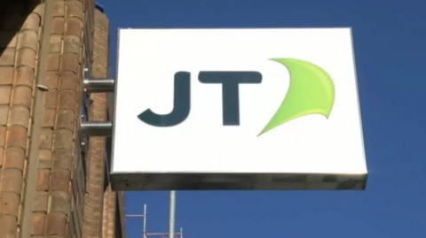 A close up of a JT Group sign against a blue sky.
