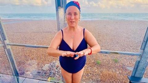 Shan wearing a blue swimsuit and a blue swim cap standing by a sea- smiling for camera.