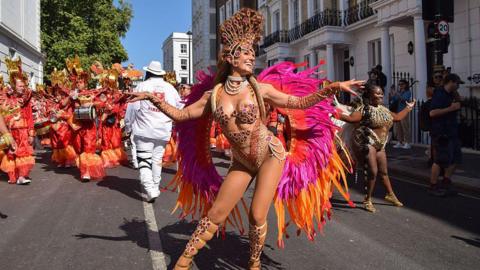 A lady in a colourful carnival dress with elaborate headdress and feathered wings poses for the cameras on a street in Notting Hill. Behind her is a troupe of performers in the same costume.
