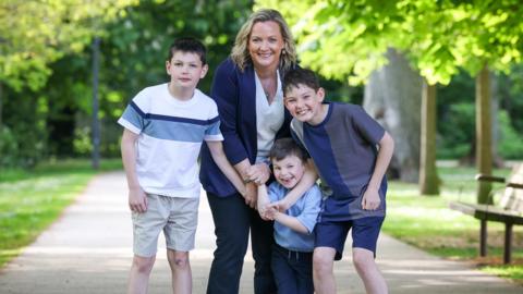 A woman in her 40s smiles in a path in a wooded area with her three sons qwho all have black hair and are also smiling 
