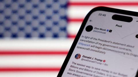 A phone in the foreground shows a social media post from Musk, about Trump; there's a US flag in the background