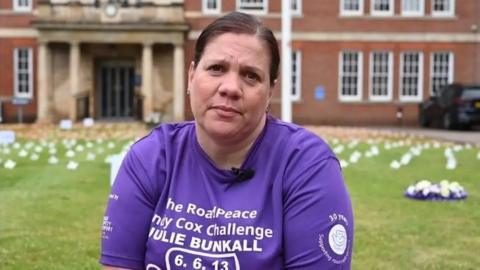 Emilie Bunkall with dark hair tied back. She is looking at the camera and wearing a purple T-shirt with white writing that says "The RoadPeace Andy Cox Challenge Julie Bunkall", together with the date of her mother's death, 6 June 2013.