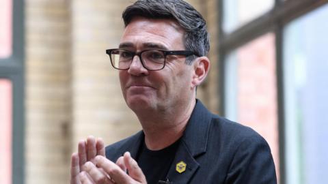 Andy Burnham clapping while wearing a dark suit and teeshirt, and a yellow broach with a bee on it.