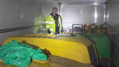 Plant machinery in the back of a lorry with a police officer dressed in high-visibility jacket. 