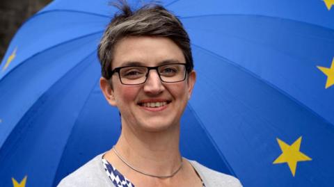 Maggie Chapman, pictured in 2019, has short hair, is wearing glasses and is smiling. Behind her is a blue umbrella with European Union yellow stars.